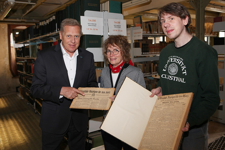 Das Foto zeigt drei Personen in einem Archiv bei der Übergabe historischer Zeitungsausgaben.